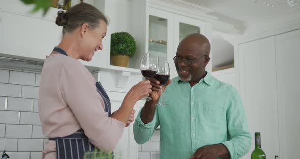 Smiling senior diverse couple wearing blue apron and drinking wine in kitchen