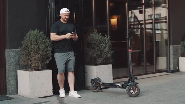 Young Man Standing with Escooter in City Downtown and Taking Selfie with Smartphone