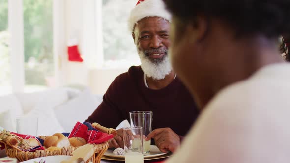 African american senior man and woman in santa hats talking and smiling while sitting on dining tabl