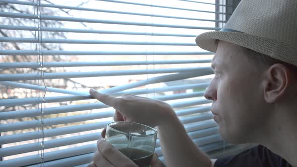 A Young Man in a Hat with Interest Examines What is Happening on the Street Drinks From a Glass