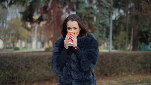 Beautiful girl drinks coffee outdoors. Portrait of a fashionable woman in fur coat with hot drink lo