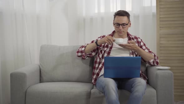 Confident Intelligent Young Man in Eyeglasses Drinking Coffee and Using Laptop Indoors. Portrait of