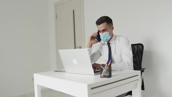 Young Student in Medical Mask Talks on a Mobile Phone When Types on a Laptop