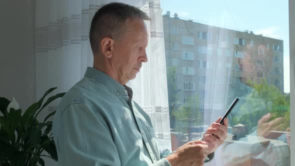 Elder Man Standing Near Window Indoors at Home While Using Mobile Phone