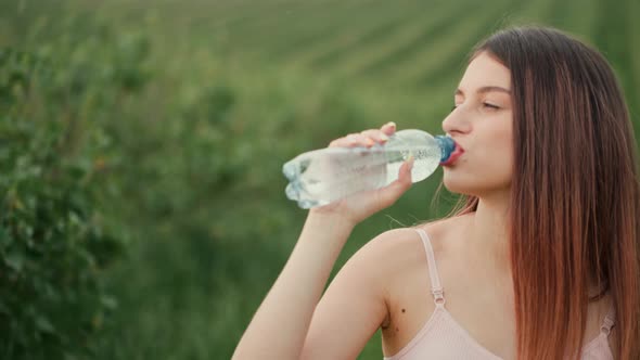 Pregnant young woman with dark hair in a pink top and light pants drinks water outdoors.