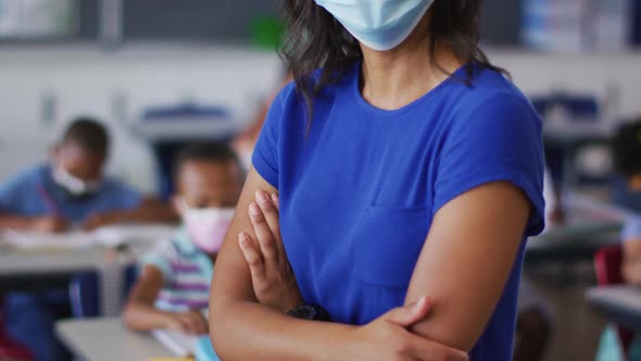 Portrait of mixed race female teacher wearing face mask, standing in classroom