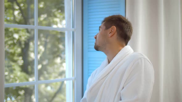 Handsome Young Bearded Man in Bathrobe Standing at Window at Home Looking Away