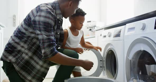 Side view of black father and son washing clothes in washing machine at comfortable home 4k