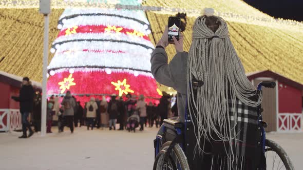 A Young Woman with Grey Dreadlocks in a Wheelchair Taking a Photo of a Bright Christmas Tree