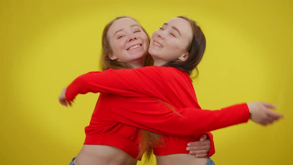 Attractive Twin Sisters Hugging Looking at Camera Posing with Toothy Smile at Yellow Background