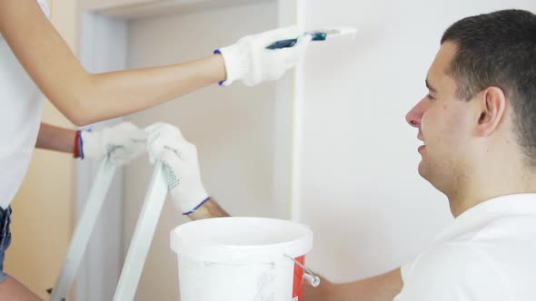 Cheerful Young Woman in White Shirt Painting the Wall in White Color Using a Brush and Standing at