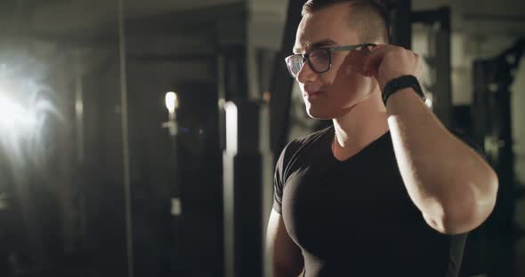 The Man in Glasses Wears Wireless Headphones on Ears in Dark Gym with Backlit