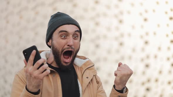 Euphoric Man Celebrating Sport Victory or Good News Using a Smart Phone in the Street Near Shopping