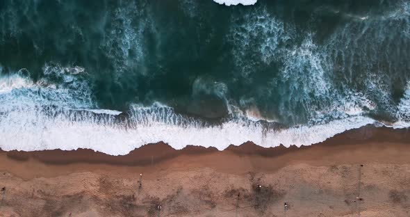 Overhead view of the waves on a beach with orange sand at sunset with people walking on the shore.