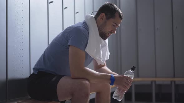 Tired Handsome Sportsman Sitting on Bench in Locker Room and Drinking Refreshing Water. Portrait of