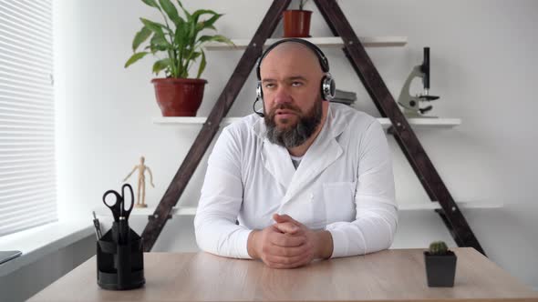 Caucasian Bearded and Bald Doctor Sits in an Office Wearing a Headset Headphones and a Microphone