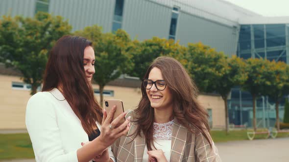 Happy Female Friends Browse the Contents of a Mobile Phone and Smile Sincerely. Two Twins Laughing