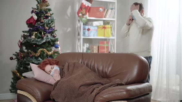 Ill Woman Lying on Armchair Coughing As Stressed Nervous Man in Covid Face Mask Talking on the Phone