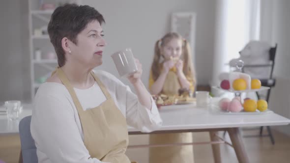 Positive Brunette Caucasian Woman Drinking Tea and Smiling at Camera As Blurred Little Girl Eating