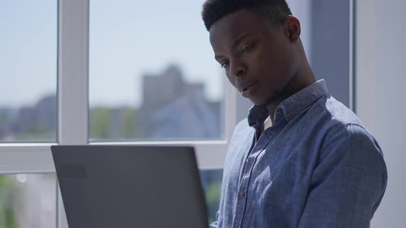 Young Concentrated Man Thinking Surfing Internet on Laptop Standing at Window in Home Office