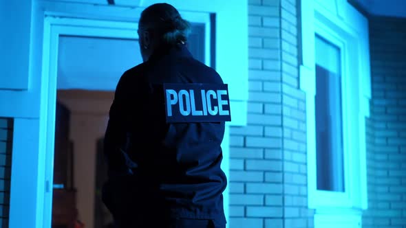 Back View Patrolman in Uniform Standing in Front of Dark House Talking on Phone As Police Lights