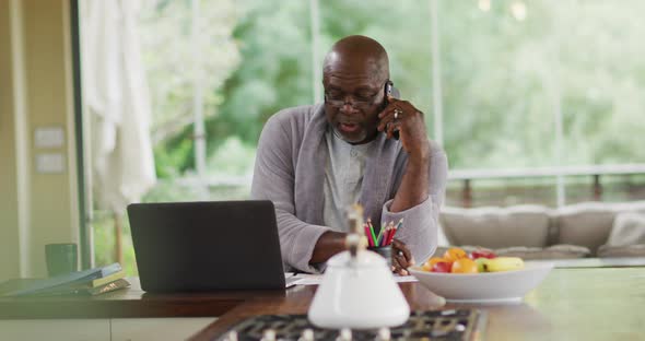 African american senior man in bathrobe sitting in kitchen using laptop and talking on smartphone