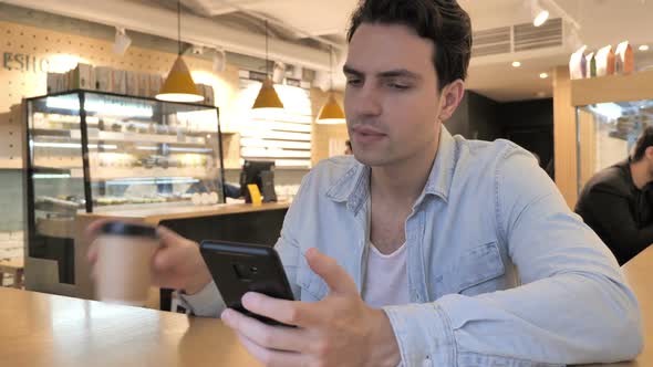 Young Man Using Smartphone and Drinking Coffee in Cafe