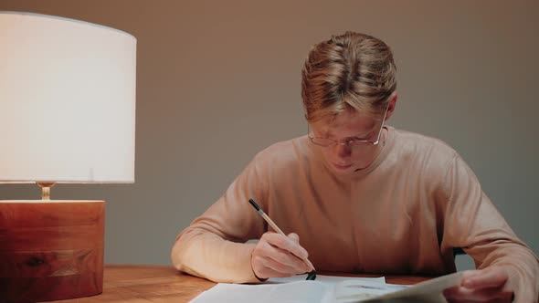 Young Man Studying at Home Writing in His Notes Reading a Textbook at the Desk