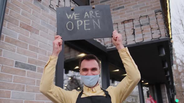 Happy Barista Holding Open Sign While Wearing Protective Face Mask and Gloves Near