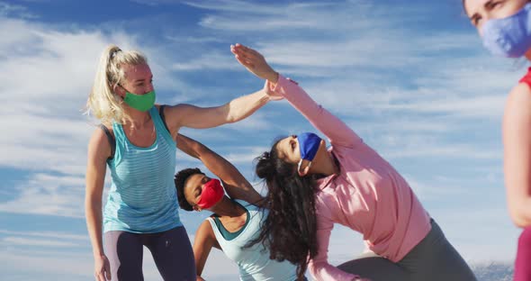 Group of diverse female friends wearing face masks practicing yoga at the beach