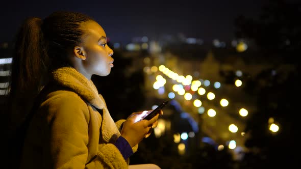 A Young Black Woman Works on a Smartphone in an Urban Area at Night  Side View