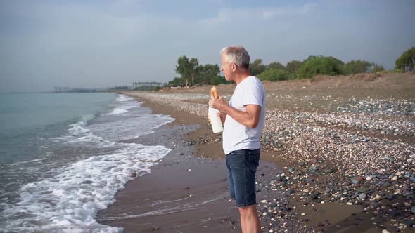 Slender Grayhaired Elderly Man Walks Barefoot on the Beach Eats Bread Washed Down with Milk From a