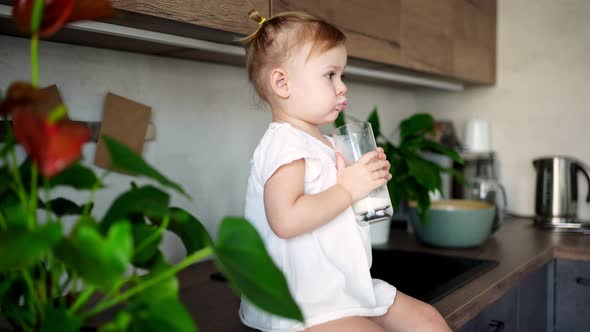 Happy Baby Girl Sitting at the Table in the Kitchen and Drinking Milk