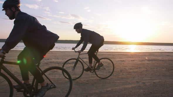 Two Young Men Riding Bicycles on the Beach on the Background of an Orange Sunsetting Sky