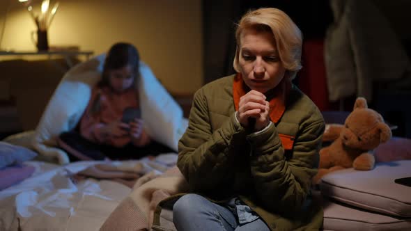 Portrait of Depressed Sad Mother Sitting on Mattress in Bomb Shelter with Blurred Daughter Playing