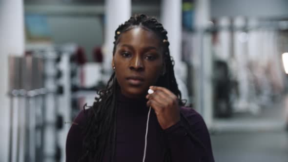 A Black Woman Walks in the Gym and Putting on Her Headphones  Looking in the Camera and Smiling