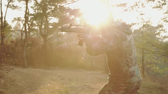 The Brutal Soldier Aims on the Object with Rifle in Forest on Dazzling Sunset