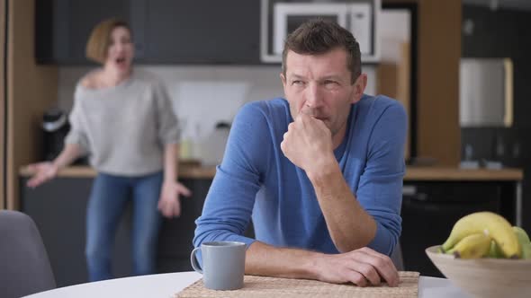 Sad Stressed Bored Man Sitting at Table Sighing As Woman Yelling at Background Arguing