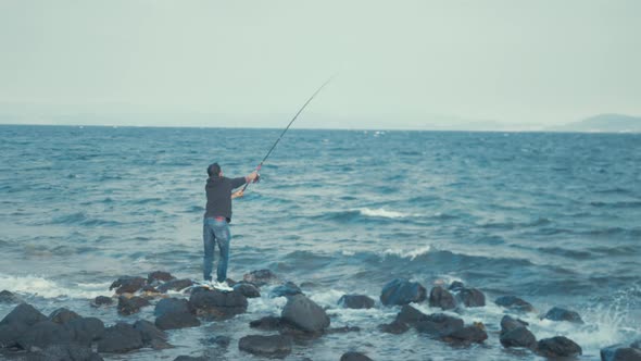 Young man casting fishing rod standing on rocks at sea shore WIDE SHOT