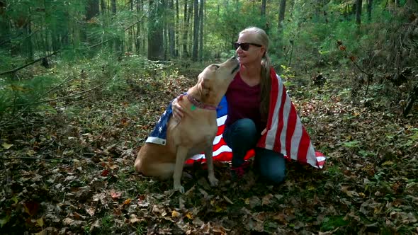 With a flag draped around them a blonde woman pets her dog and the dog looks up.