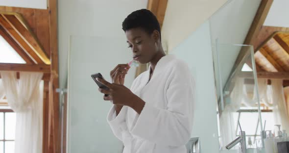 African american attractive woman brushing teeth and using smartphone in bathroom
