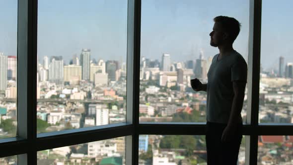 Dreaming European Man Stands By the Window with Great Views and Drinks Hot Coffee or Tea Enjoying