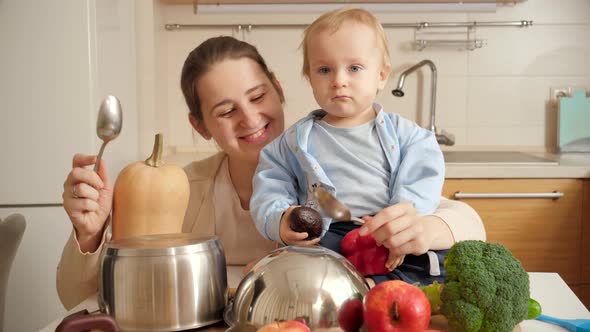 Happy Smiling Mother with Baby Son Playing with Spoons on Pans in Kitchen Like on Drums