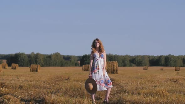 Beautiful Redhaired Woman in a White Dress and Hat Stands on the Background of the Field