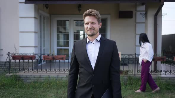 Confident Smiling Man Gesturing Yes Walking on Front Yard with Blurred Woman Strolling at Background