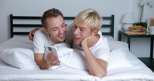 Young Gay Couple Lying on Bed in Bedroom and Using Smartphone Together