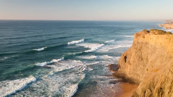 Aerial drone shot of waves hitting a beach with cliffs during a sunset in southern Portugal