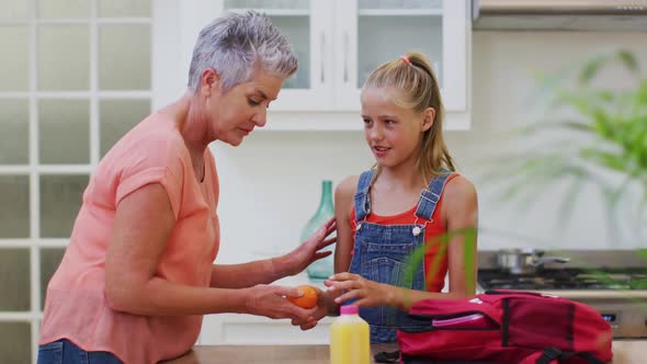 Caucasian grandmother in kitchen preparing packed lunch talking with granddaughter giving her fruit