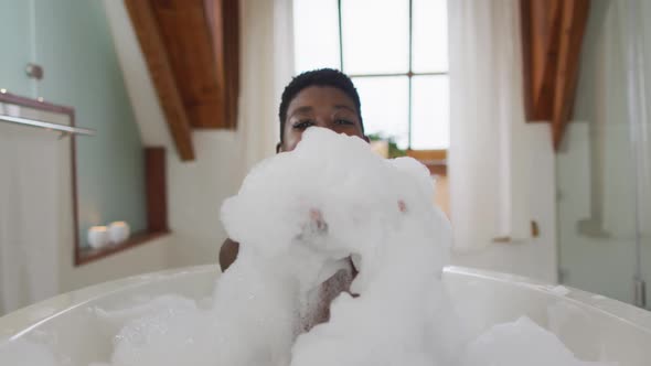 Smiling african american attractive woman taking bath and blowing out foam in bathroom