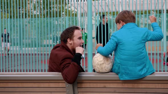 Young Man and His Little Brother Sitting on the Bench and Watching Other Kids Playing Football